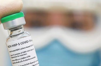 person wearing face mask and safety goggles holds up labelled injection vial with gloved hand