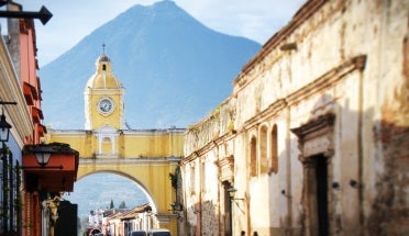 Rows of colorful buildings line the street leading to the famous yellow Saint Catalina archway in historic Antigua Guatemala with Agua volcano looming above in the distance.